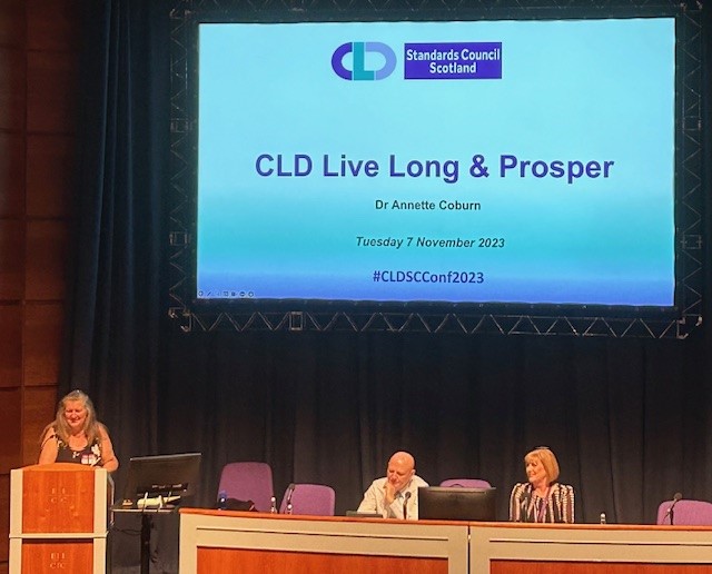 Dr Annette Coburn standing at a lecturn with the title slide of her presentation on a screen behind her.  It has the CLD Standards Council logo at the top and underneath the title CLD Live Long and Prosper.  To the right of the screen, Alan Sherry and Dr Marion Allison are sitting at the top table.